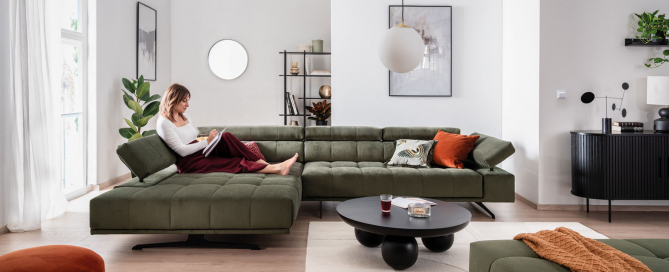 Frau entspannt auf dem Sofa Raimo in dunkelgrünem Stoff in einem hellen Wohnzimmer