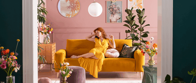 Frau sitzt im gelben Kleid auf dem gelben Sofa Alika in einem frühlingshaft dekorierten Zimmer